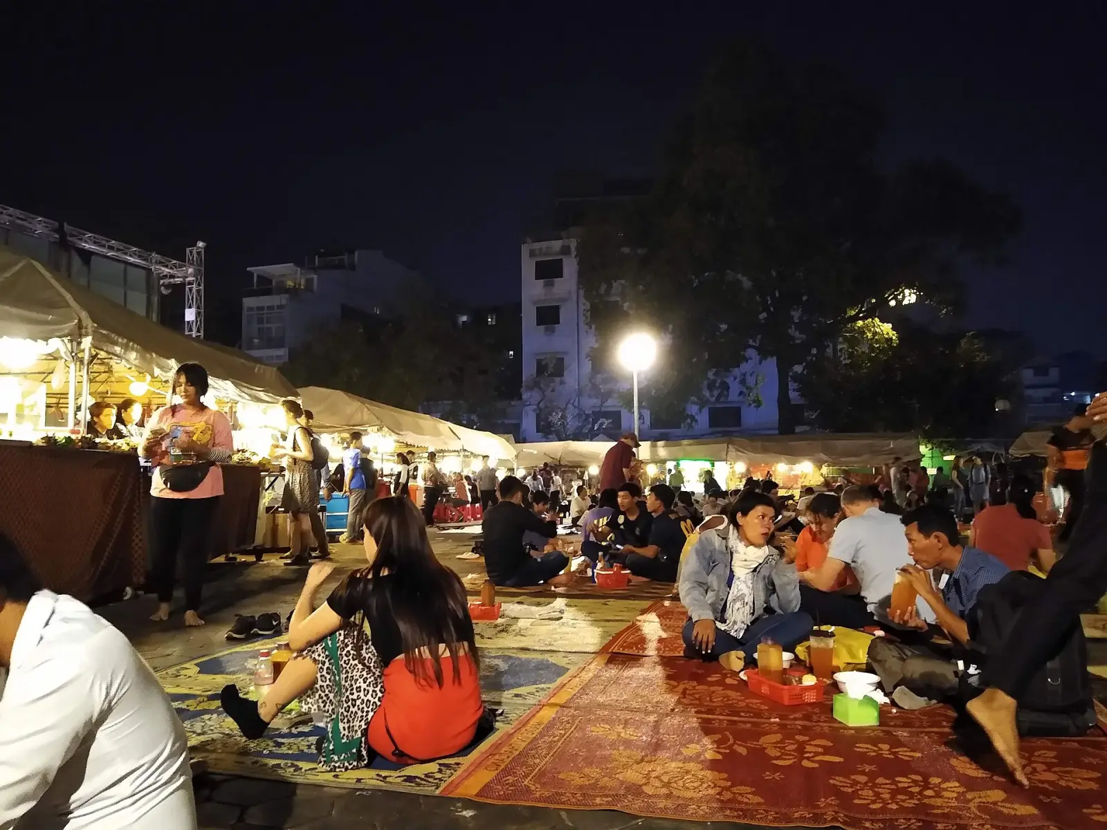 A Phnom Penh night market after dark, with lit food stalls behind people eating on woven mats