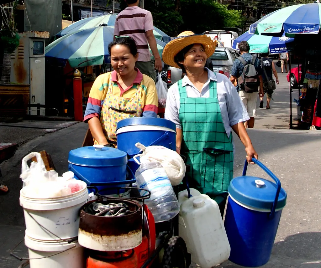 Two street food vendors in aprons pushing carts loaded with buckets and containers along a Bangkok street
