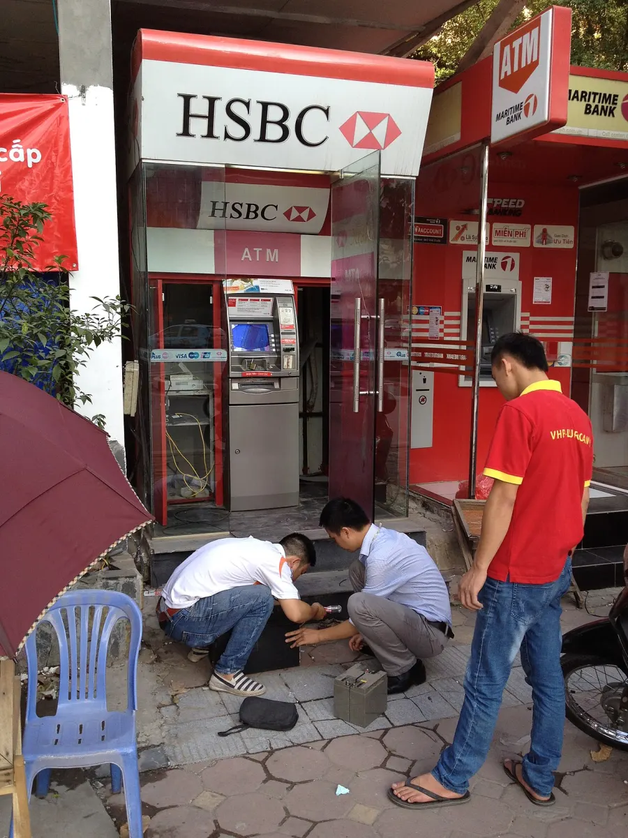 Two street-side ATM booths in Hanoi, one HSBC and one Maritime Bank, with technicians working at the kerb